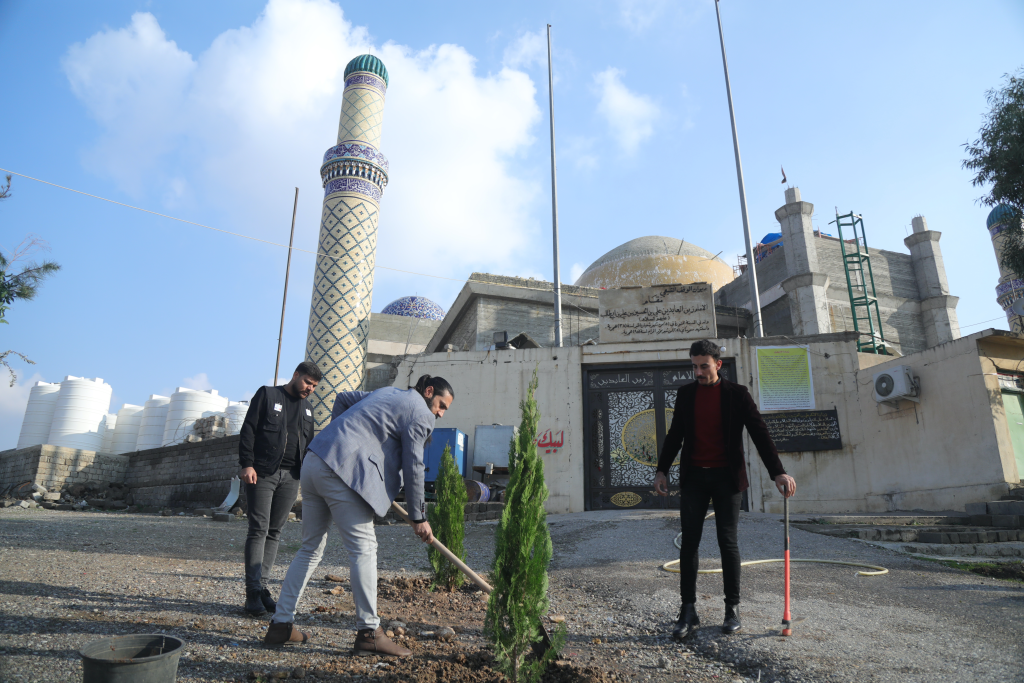 Three men plant trees in front of the Imam Zain Al-Abidin Shrine.