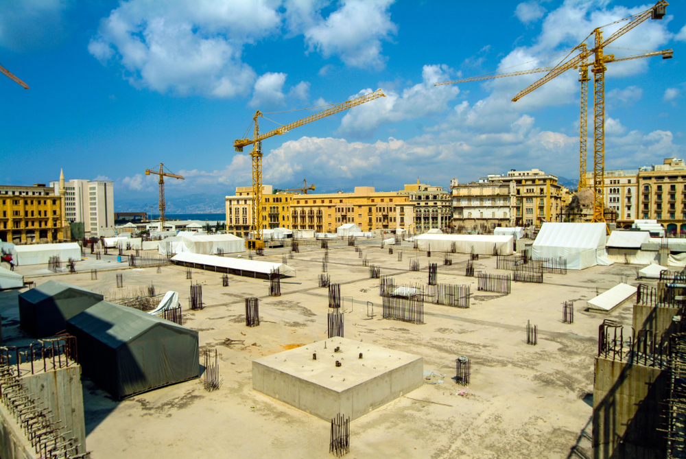 A building site: empty land with three tall cranes and buildings in the background