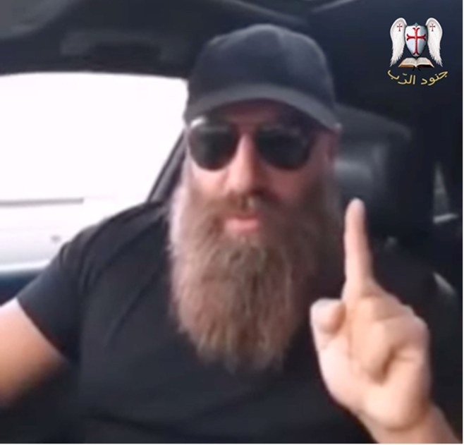 A man sat in a car wearing a cap and sunglasses and holding one finger up